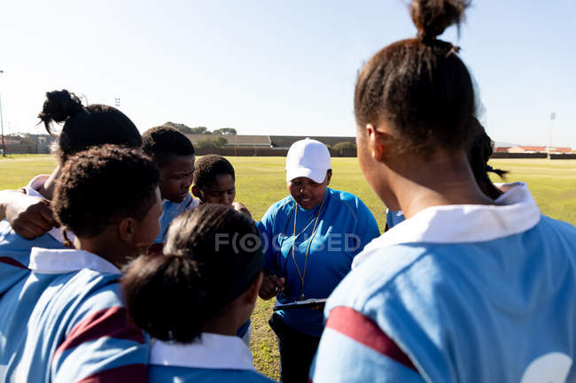 Rear view of a team of young adult multi-ethnic female rugby players standing on a rugby field with arms linked listening to their middle aged mixed race female coach while preparing for a rugby match — Stock Photo