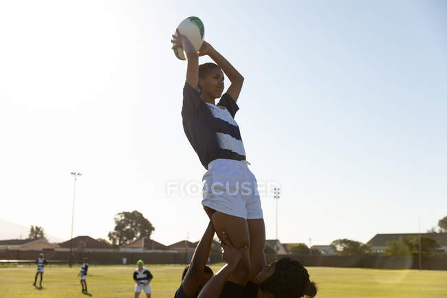 Vista lateral de uma jovem adulta mista jogador de rugby feminino sendo levantado por companheiros de equipe para pegar a bola durante uma partida de rugby — Fotografia de Stock