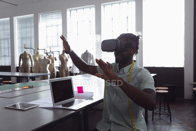 Front view of a young African American male fashion student with his hands raised using a VR headset in a studio at fashion college with a computer and a tablet computer on the table in front of him, and mannequins in the background — Stock Photo