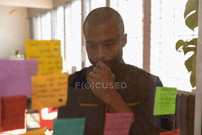 Front view close up of a young African American man reading notes on a glass wall and thinking during a team brainstorm session at a creative office, seen through glass wall — Stock Photo