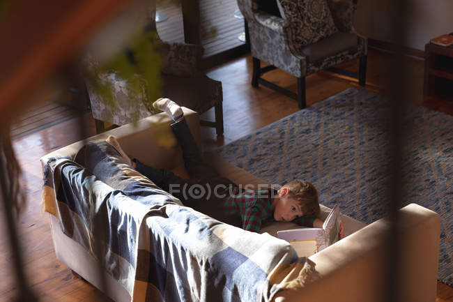 Vista lateral de un niño caucásico preadolescente acostado en un sofá en una sala de estar y leyendo un libro - foto de stock