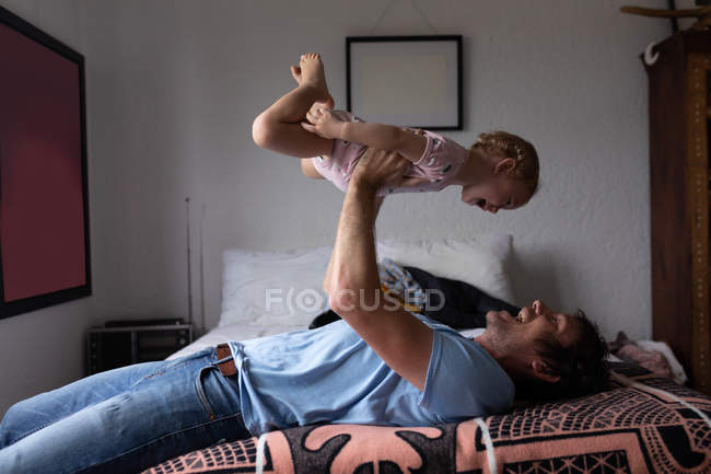 Вид сбоку на молодого кавказского отца, держащего своего ребенка над собой и лежащего на кровати — стоковое фото