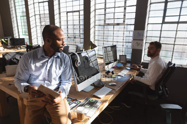 Front view close up of a young African American man sitting on a desk using a tablet computer and talking with a young Caucasian male colleague sitting at a desk using a computer by a window in a creative office — Stock Photo