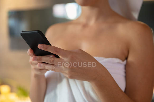 Sección media de una mujer que usa una toalla de baño usando un teléfono inteligente en un baño - foto de stock