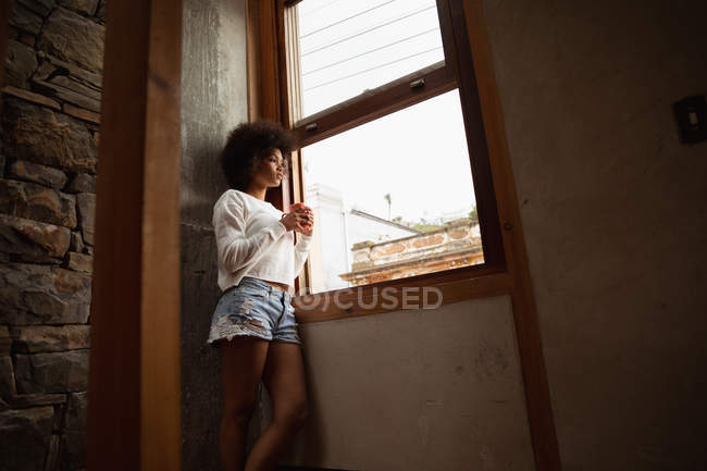 Vista lateral de uma jovem mulher de raça mista em pé perto de uma janela e olhando para fora segurando uma xícara de café — Fotografia de Stock