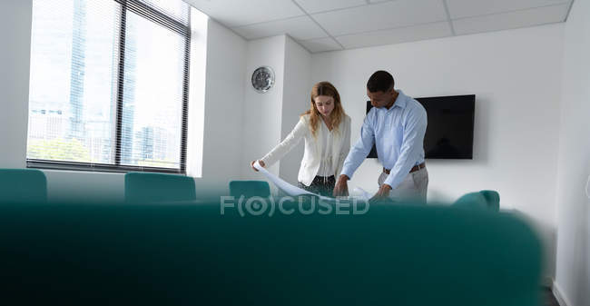 Vue de face d'un jeune homme afro-américain et d'une jeune femme caucasienne debout et parlant à un bureau tenant un dessin de plan dans le bureau moderne d'une entreprise créative — Photo de stock