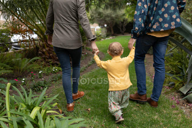 Вид сзади на низкую часть молодых кавказских отцов и матерей, держащихся за руки с малышом и гуляющих в саду — стоковое фото