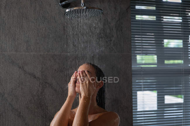 Gros plan vue de face d'une jeune femme brune caucasienne debout sous la douche avec les mains sur les yeux dans une salle de bain moderne — Photo de stock