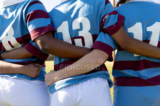Vista lateral de uma equipe de jogadores de rugby femininos em pé em um campo de rugby com os braços ligados se preparando para uma partida de rugby — Fotografia de Stock