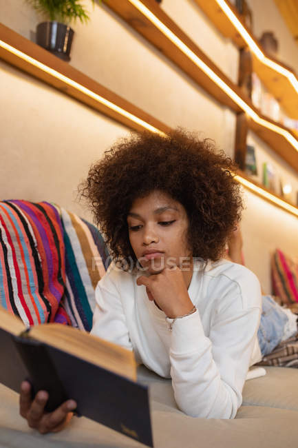 Vista frontal close up de uma jovem mulher de raça mista deitada em sua frente em um sofá lendo um livro em casa, apoiando-se em seu cotovelo com o queixo descansando em sua mão — Fotografia de Stock