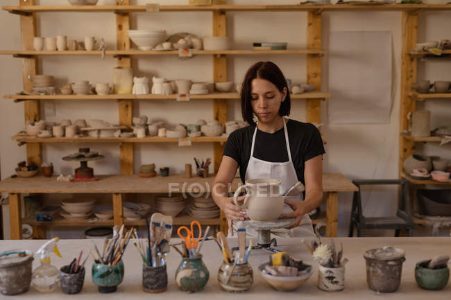 Front view of a young Caucasian female potter standing at a work table holding a jug on a banding wheel in a pottery studio, with pots and tools in the foreground — Stock Photo