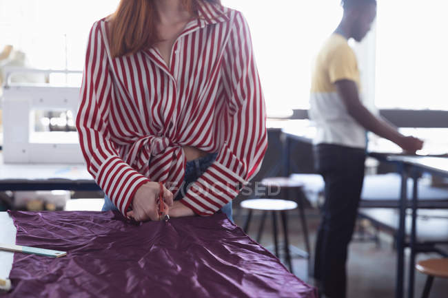Front view mid section of a young Caucasian female fashion student cutting purple fabric while working on a design in a studio at fashion college, with a student working in the background — Stock Photo