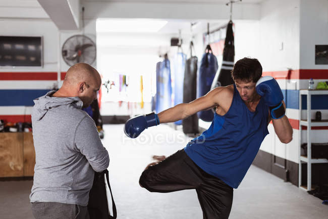 Vue de face d'un jeune boxeur masculin caucasien pratiquant un coup de pied contre un pad tenu par un entraîneur masculin caucasien d'âge moyen dans un gymnase de boxe — Photo de stock