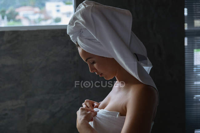 Vista lateral de cerca de una joven mujer caucásica con una toalla de baño y con el pelo envuelto en una toalla, de pie en un baño moderno mirando hacia abajo - foto de stock