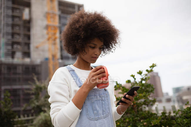 Vista laterale da vicino di una giovane donna di razza mista in piedi fuori su un balcone della città con una tazza di caffè e guardando il suo smartphone — Foto stock