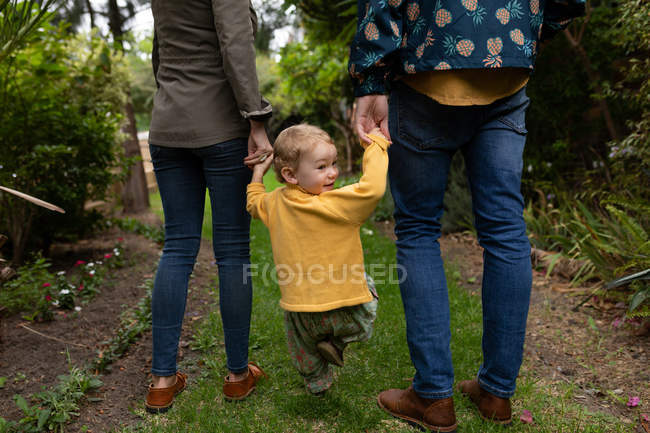 Vista trasera de una sección baja de un joven padre y madre caucásicos tomados de la mano con su bebé y caminando en un jardín, el bebé está dando la vuelta - foto de stock