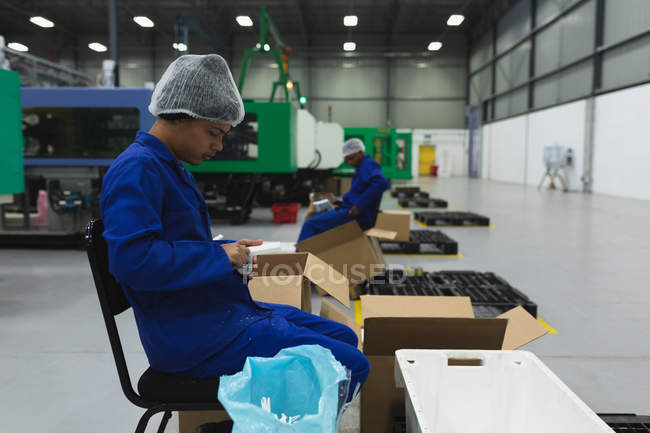Side view close up of a young mixed race man sitting and packing product into boxes for shipping in a warehouse at a processing plant, a coworker visible in the background — Stock Photo