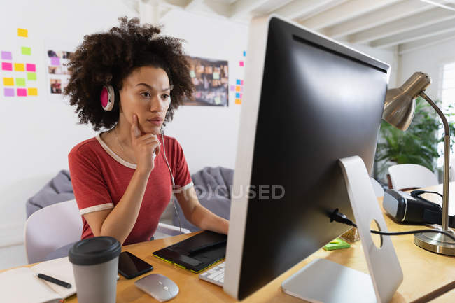 Seitenansicht einer jungen Frau mit gemischter Rasse, die mit Kopfhörern an einem Schreibtisch sitzt und auf einen Computerbildschirm in einem Kreativbüro blickt — Stockfoto