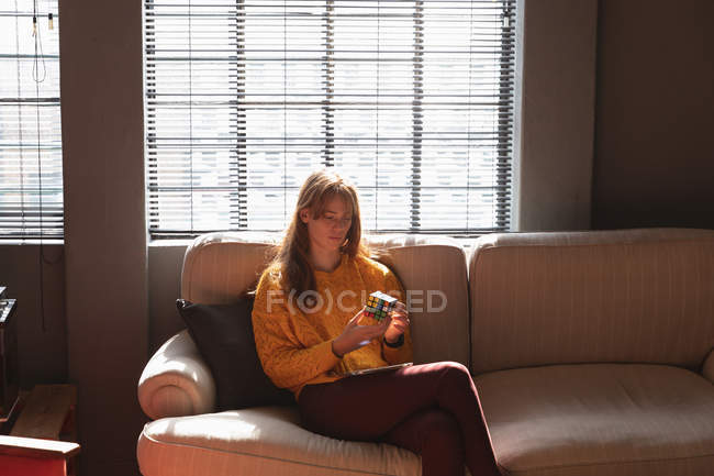 Vista frontal de uma jovem caucasiana sentada em um sofá usando um smartphone na área de estar de um escritório criativo, iluminada pela luz solar — Fotografia de Stock