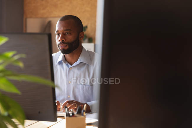 Vista frontal de cerca de un joven afroamericano sentado en un escritorio usando una computadora en una oficina creativa, vista entre pantallas de computadora - foto de stock