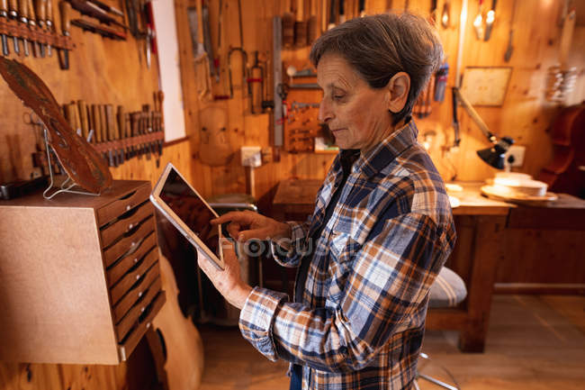 Side view of a senior Caucasian female luthier using a tablet computer with tools hanging up on the wall in the background — Stock Photo