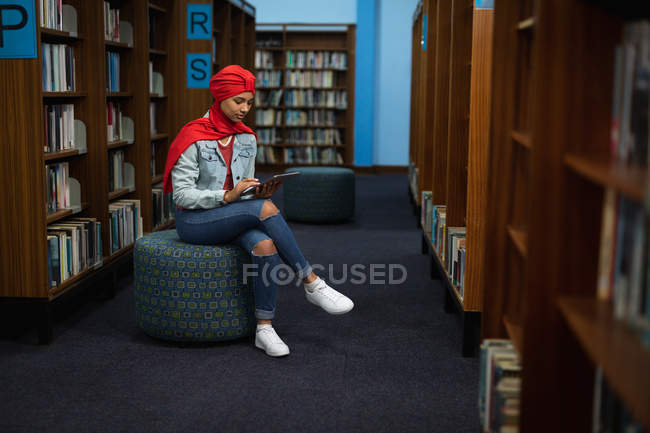 Вид сбоку на молодую азиатскую студентку в тюрбане, которая пользуется планшетным компьютером и учится в библиотеке — стоковое фото