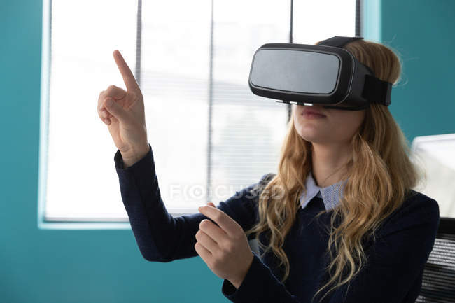 Front view close up of a young Caucasian woman sitting wearing a VR headset and with one hand raised and finger pointing in the modern office of a creative business — Stock Photo
