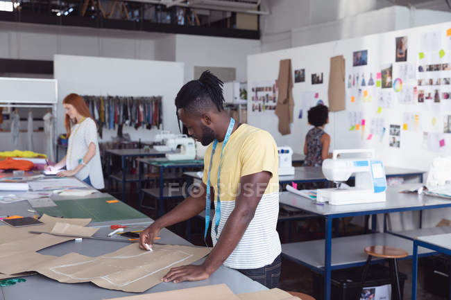 Vista laterale di un giovane afroamericano caucasico studente di moda maschile che disegna un modello mentre lavora su un design in uno studio al college di moda, con altri studenti che lavorano sullo sfondo — Foto stock