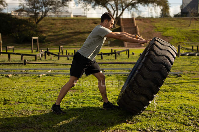 Vista lateral de un joven caucásico volteando un neumático en un gimnasio al aire libre durante una sesión de entrenamiento de bootcamp - foto de stock