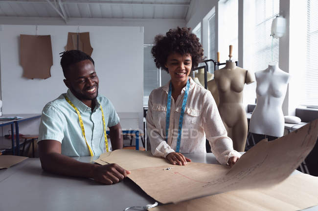 Вид спереди на молодого афроамериканского мужчину и молодую студентку смешанной расы, держащую шаблон, работая над дизайном в студии в колледже моды, с манекенами на заднем плане — стоковое фото
