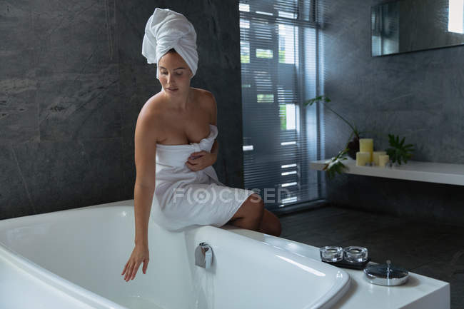 Vista frontal de una joven mujer caucásica con una toalla de baño y con el pelo envuelto en una toalla, sentada en el borde de la bañera y tocando el agua en un baño moderno - foto de stock
