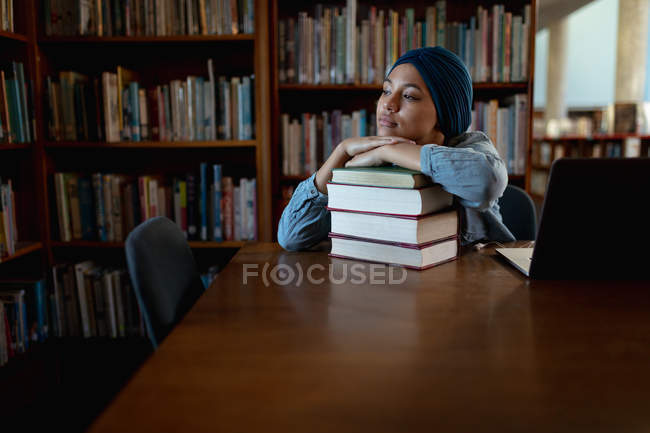 Vista frontal de cerca de una joven estudiante asiática vestida con un turbante descansando sobre una pila de libros y estudiando en una biblioteca - foto de stock