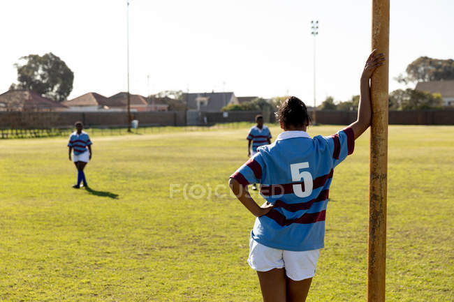 Rückansicht einer jungen erwachsenen gemischten Rugby-Spielerin, die auf einem Rugbyfeld an einen Torpfosten gelehnt steht, mit ihren Teamkolleginnen im Hintergrund — Stockfoto