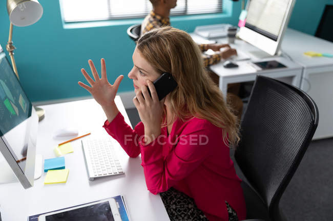 Vista lateral de cerca a una joven mujer caucásica sentada en un escritorio frente a una computadora hablando en un teléfono inteligente y haciendo un gesto con la mano levantada - foto de stock