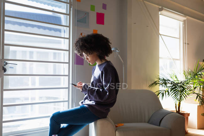 Side view of a young mixed race woman sitting on an armchair and using a smartphone at a creative office — Stock Photo