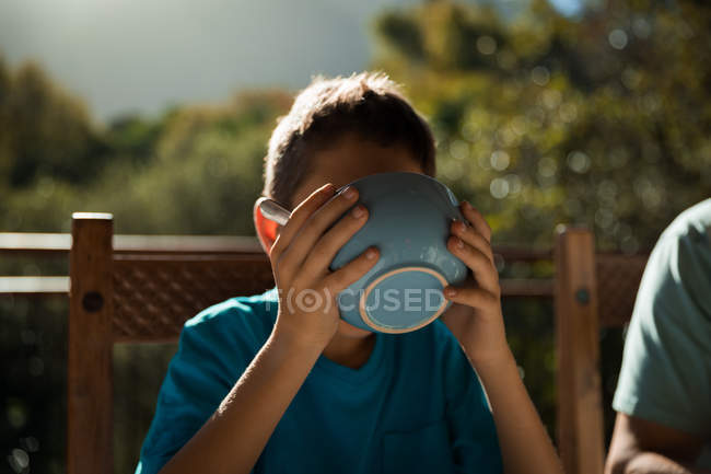 Vista frontal de cerca de un niño caucásico pre adolescente sentado en una mesa disfrutando del desayuno en un jardín, bebiendo de un tazón - foto de stock