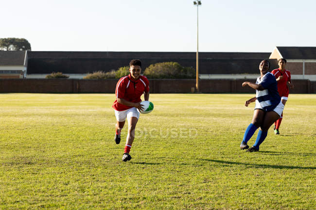 Frontansicht von drei jungen erwachsenen gemischten Rugbyspielerinnen, die während eines Rugbyspiels laufen, wobei ein Spieler den Ball hält — Stockfoto