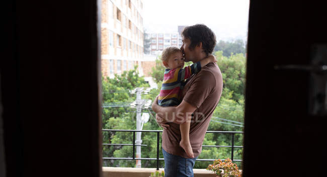 Seitenansicht eines jungen kaukasischen Vaters mit seinem Baby auf einem Balkon mit einem Park im Hintergrund — Stockfoto