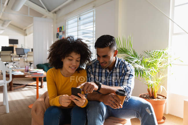 Вид спереди на молодого человека смешанной расы и молодую расистку, сидящую на бобовом мешке с помощью смартфона и улыбающуюся в креативном офисе — стоковое фото