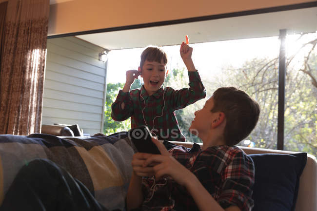 Вид спереди на двух мальчиков дошкольного возраста, использующих смартфон и слушающих музыку с включенными наушниками в гостиной дома — стоковое фото