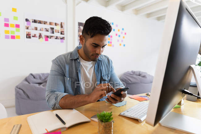 Side view of a young mixed race man sitting at a desk and using a smartphone at a creative office — Stock Photo