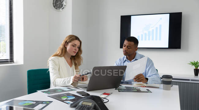 Vue de face d'un jeune homme afro-américain et d'une jeune femme caucasienne assis et parlant à un bureau à l'aide d'un ordinateur portable dans le bureau moderne d'une entreprise créative — Photo de stock