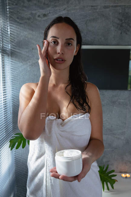 Portrait close up of a young Caucasian brunette woman wearing a bath towel applying face cream in a modern bathroom — Stock Photo