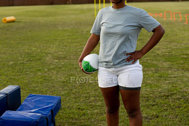 Vista frontal sección media de la jugadora de rugby de pie en un campo de deportes con la mano en la cadera sosteniendo una pelota de rugby durante una sesión de entrenamiento - foto de stock