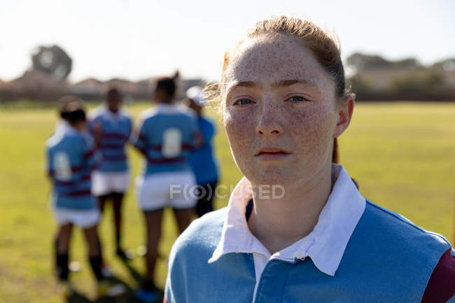 Portrait close up of a young adult Caucasian female rugby player standing on a rugby pitch looking to camera, with her teammates talking together in the background — Stock Photo