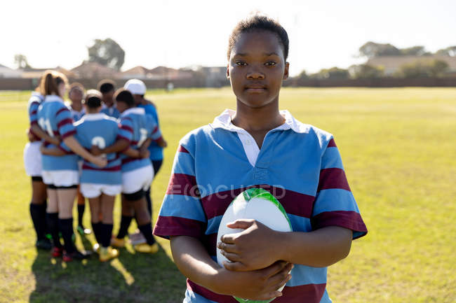 Portrait of a young adult mixed race female rugby player standing on a rugby pitch holding a rugby ball in her hands looking to camera, with her teammates in a huddle together in the background — Stock Photo