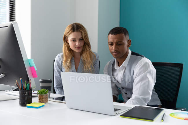 Vista frontal de cerca de un joven afroamericano y una joven mujer caucásica usando un ordenador portátil juntos sentados en un escritorio en la oficina moderna de un negocio creativo - foto de stock