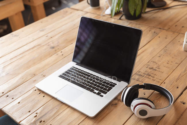 Gros plan d'un ordinateur portable et d'une paire d'écouteurs assis sur un bureau en bois dans un espace de bureau créatif — Photo de stock
