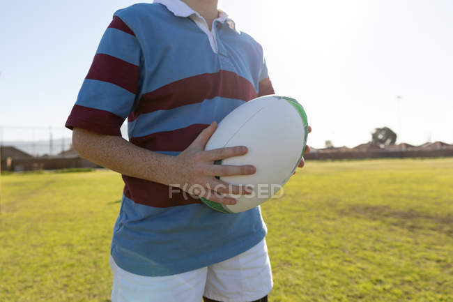 Vorderseite Mittelteil einer Rugbyspielerin, die auf einem Rugbyfeld steht und einen Rugbyball unter ihrem Arm hält — Stockfoto
