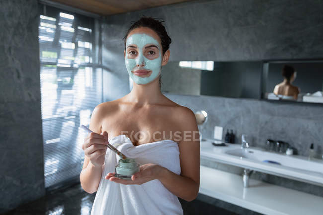 Retrato de una joven mujer caucásica con una toalla de baño sosteniendo un frasco de paquete facial y sumergiéndose en un cepillo en preparación para aplicarlo en su cara, mirando a la cámara sonriendo - foto de stock